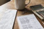 Grocery receipt, notepad of crossed-out monthly expenses, calculator, and coins on a wooden table beside a coffee mug